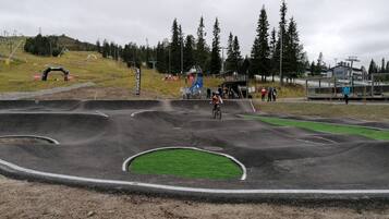 A BMX track with a cyclist in action, surrounded by spectators and a hilly landscape with trees and a ski lift in the background.