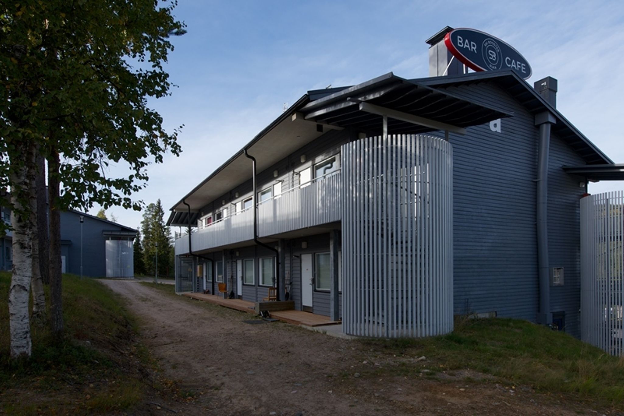 Un bâtiment moderne, avec une enseigne indiquant la présence d’un bar et d’un café, une allée couverte et un chemin de terre menant à l’entrée.