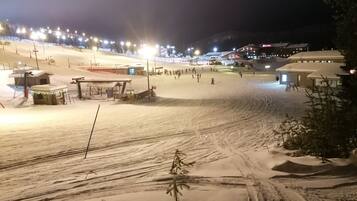 A snow-covered ski slope at night with illuminated lights, ski lifts, and buildings in the background.