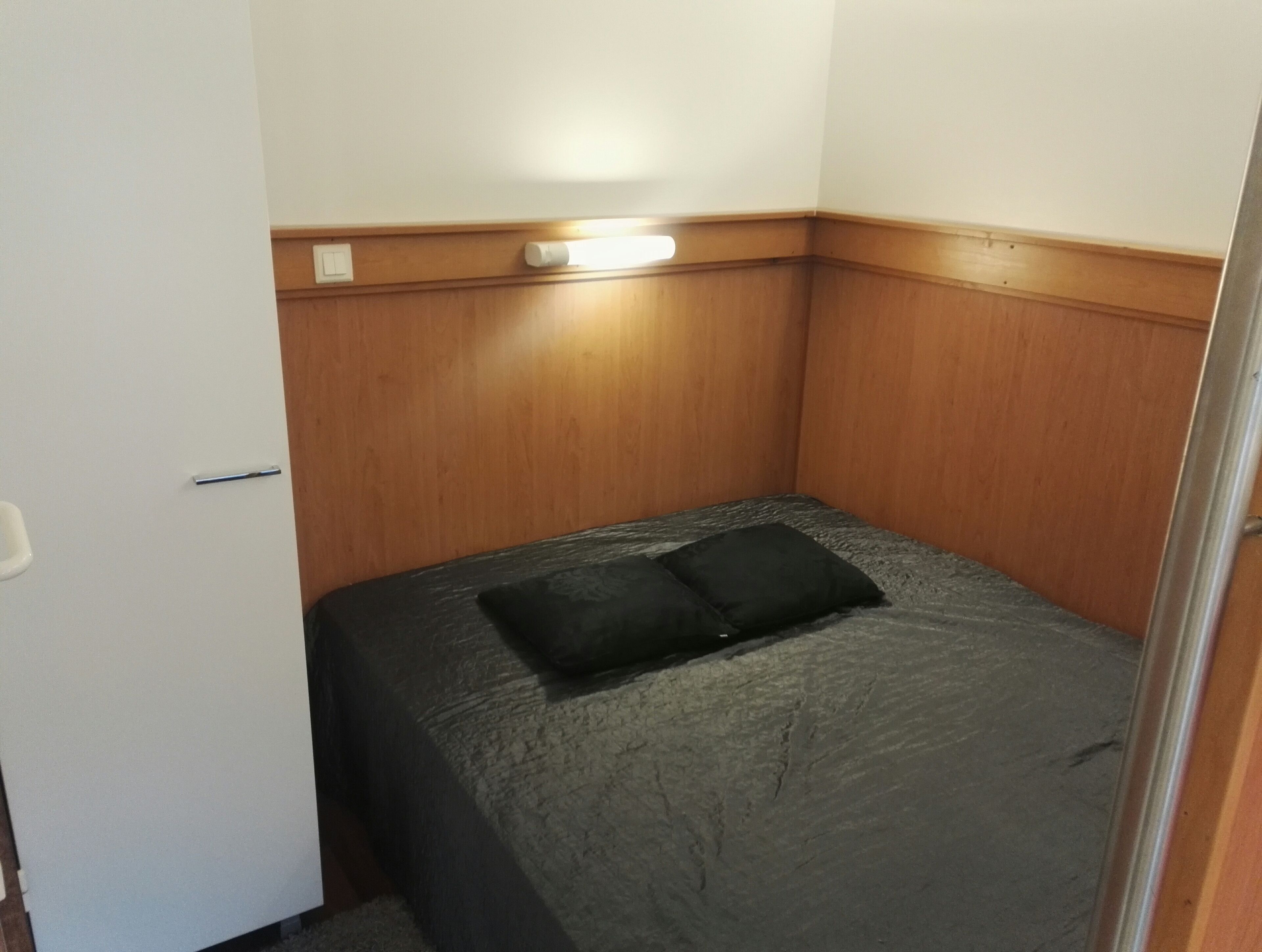 A small, narrow room with a single bed, a wooden panel wall, and a light fixture above the bed.