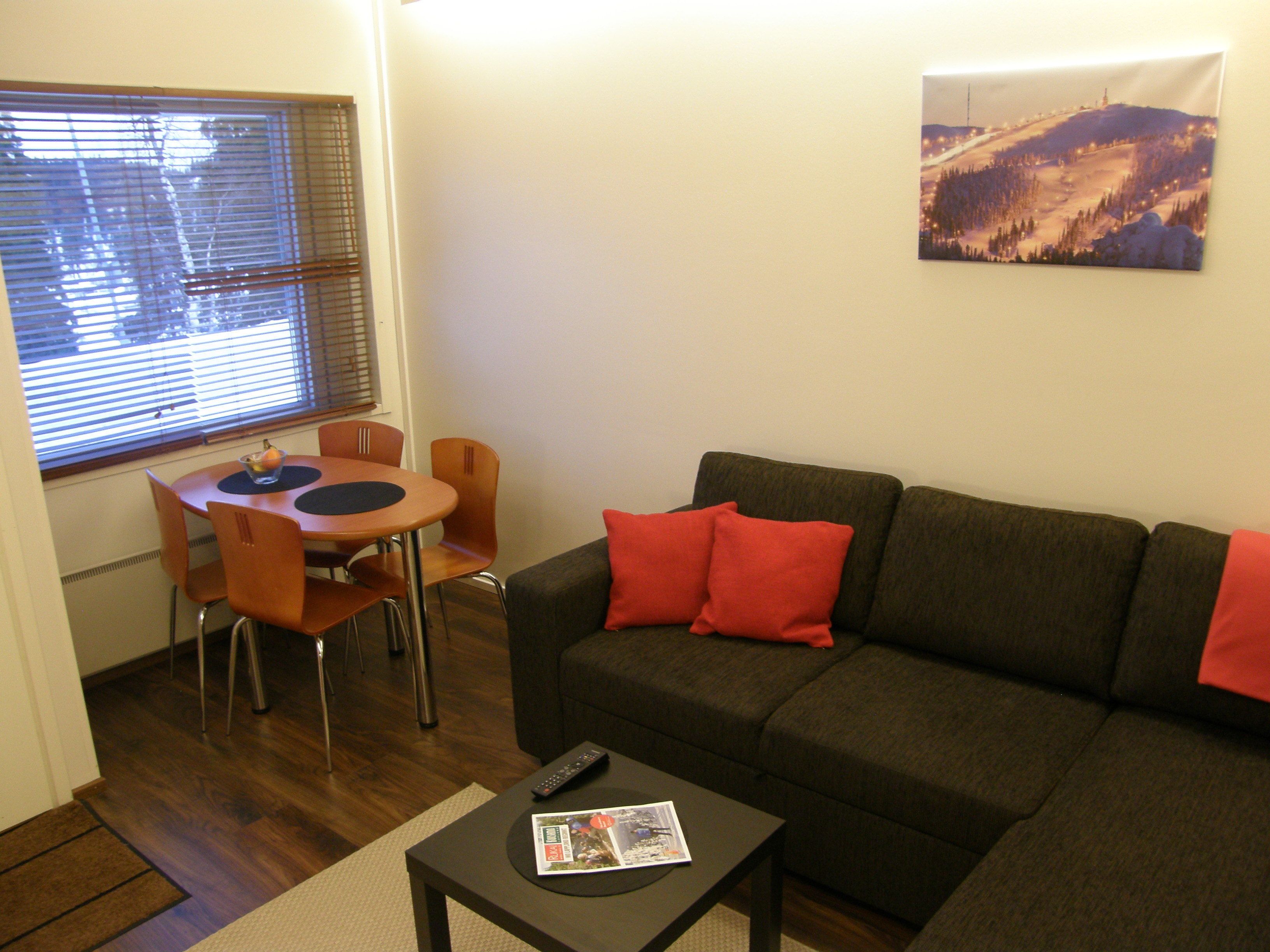 A living room with a dark grey sofa, two red cushions, a small round table with a remote control and a magazine, and a window with blinds.