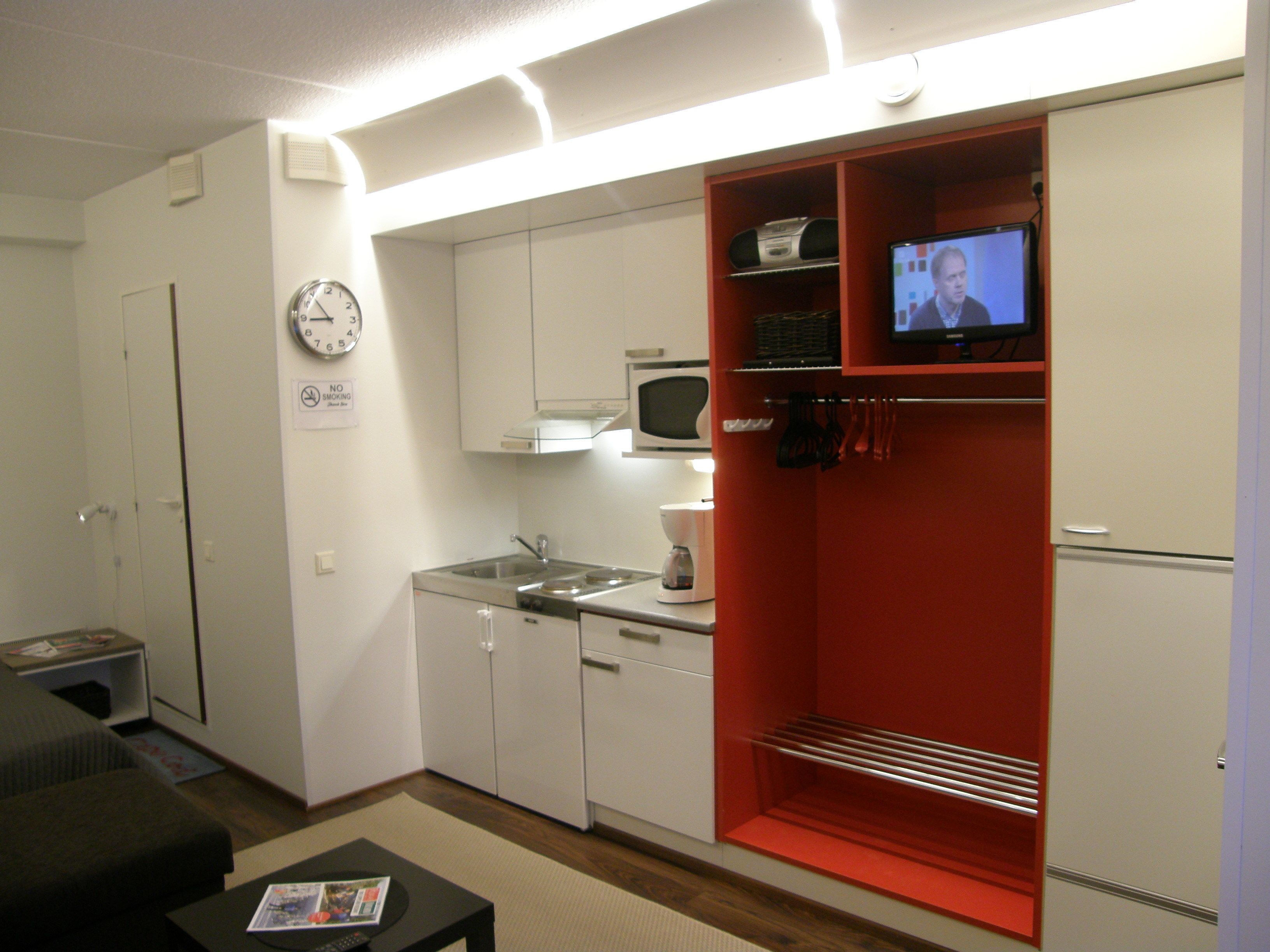 A compact hotel room with a kitchenette, a television, and a clock on the wall.