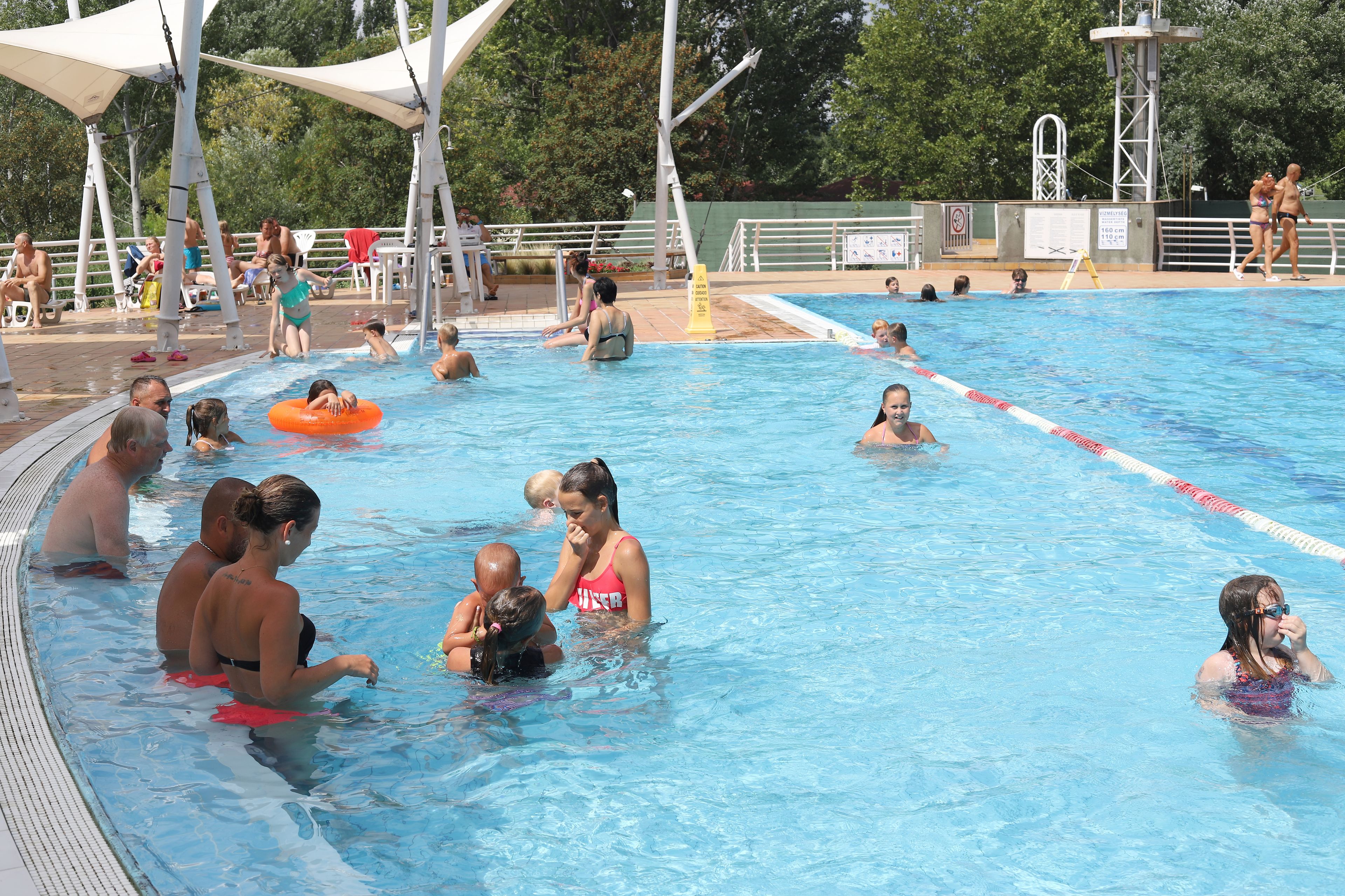 A swimming pool with people in the water, some with floatation devices, and others sitting or standing on the poolside.