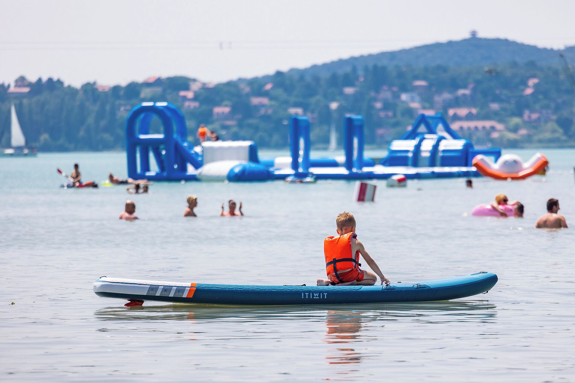 Eine Person sitzt auf einer blauen Luftmatratze im Wasser, im Hintergrund sind aufblasbare Objekte zu sehen.