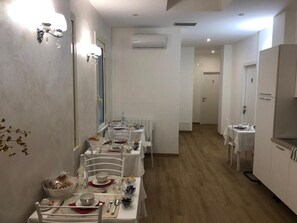 Un coin repas avec des tables et des chaises blanches, un espace cuisine équipé d’un réfrigérateur, et un appareil de climatisation fixé au mur.