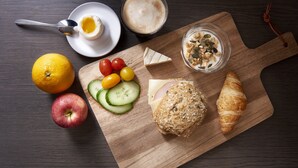 Una tabla de madera con rodajas de pepino, tomates cherry, un croissant, una loncha de queso y un vaso de leche con granola sobre una mesa de madera oscura.