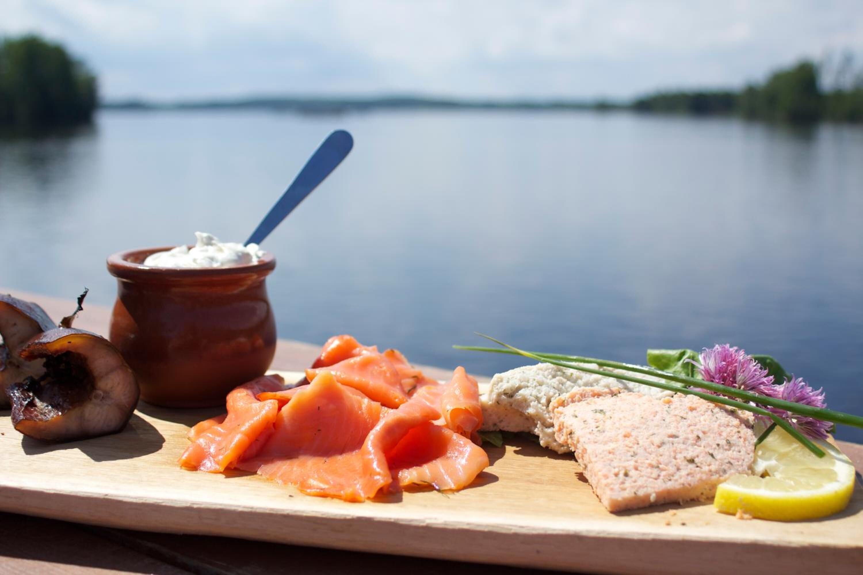 Un tagliere di legno con diversi alimenti tra cui salmone affumicato, una crema spalmabile e una fetta di carne, sullo sfondo di un lago calmo.