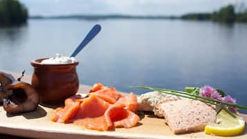 Un tagliere di legno con diversi alimenti tra cui salmone affumicato, una crema spalmabile e una fetta di carne, sullo sfondo di un lago calmo.
