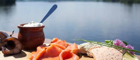 Une planche en bois sur laquelle sont disposés divers aliments, notamment du saumon fumé, une pâte à tartiner crémeuse et une tranche de viande, le tout présenté sur fond d’un lac paisible.
