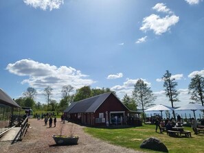 A rustic barn-style building with a gravel path leading to it, surrounded by trees and picnic tables.