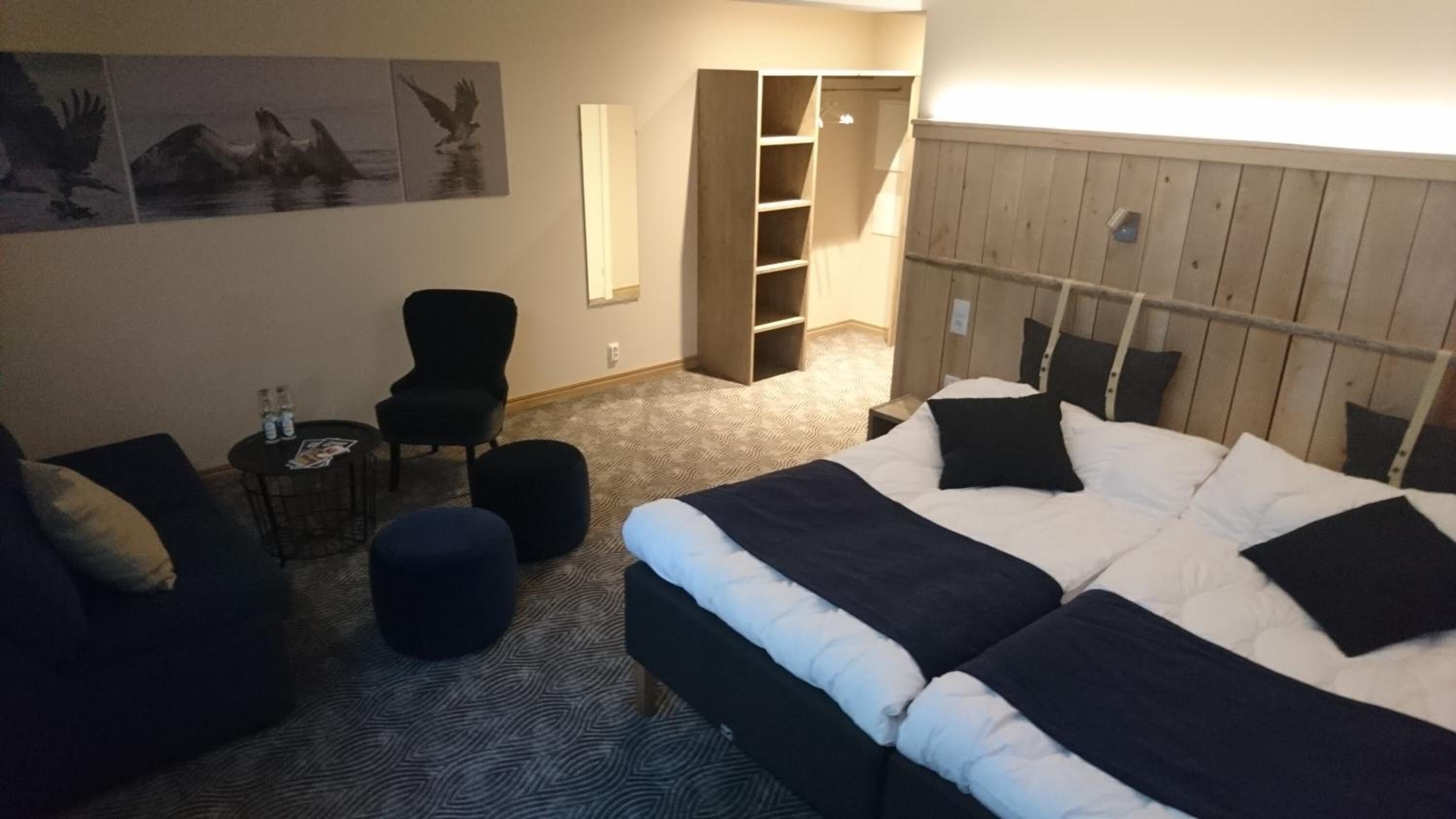 Una habitación de hotel con una cama grande, una silla, una mesita con botellas y una estantería.