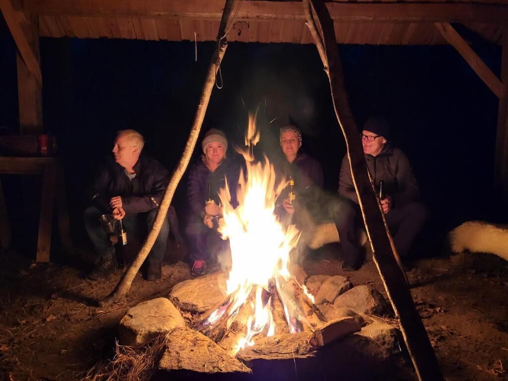Vier Personen sitzen an einem Lagerfeuer in einer rustikalen Naturlandschaft.