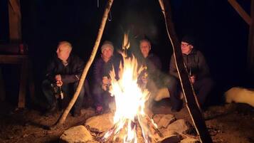 Vier Personen sitzen an einem Lagerfeuer in einer rustikalen Naturlandschaft.