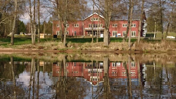A large, red brick building with a symmetrical facade, surrounded by trees and reflecting in a calm body of water.