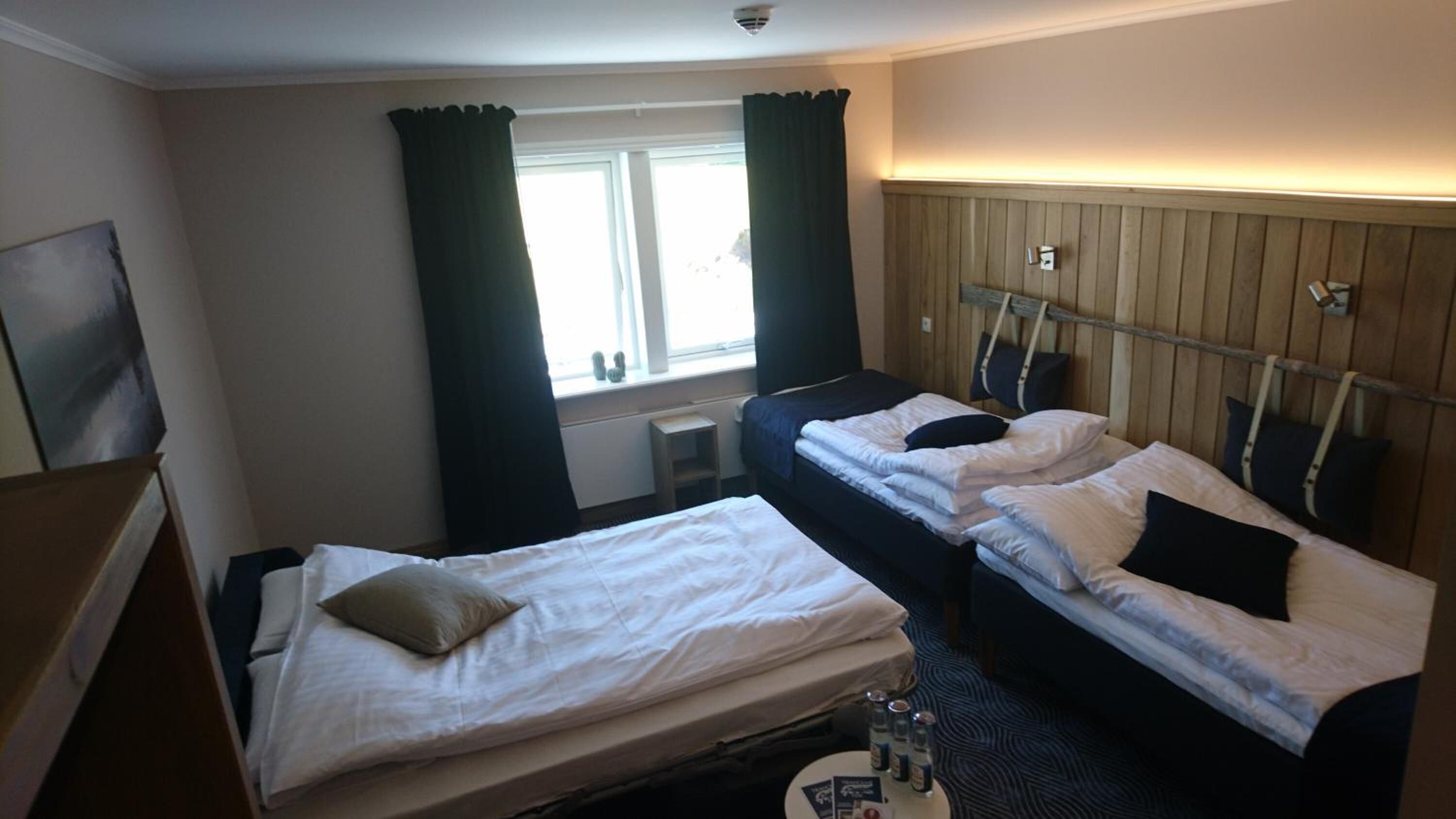 A hotel room with two single beds, a small table with a bottle, and a window with curtains.
