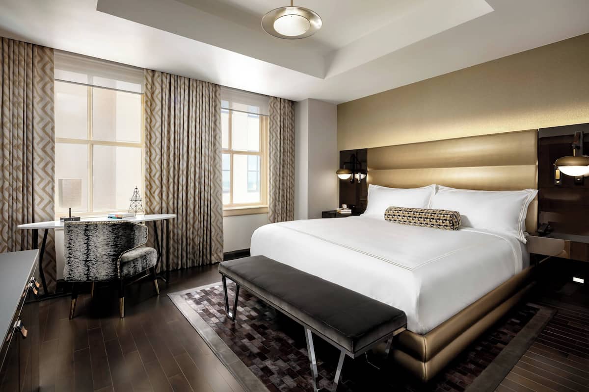 A hotel room with a large bed, a desk with a chair, a bench, and a view of the city through the window.