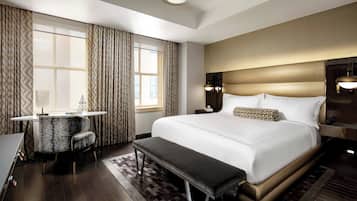A hotel room with a large bed, a desk with a chair, a bench, and a view of the city through the window.