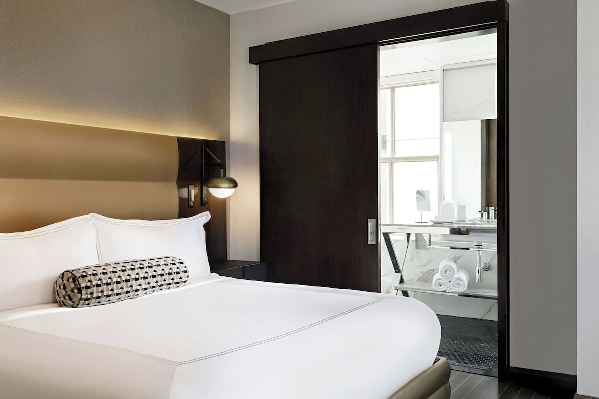 A modern hotel room with a large bed, a dark wooden door, and a bathroom visible through a glass door.