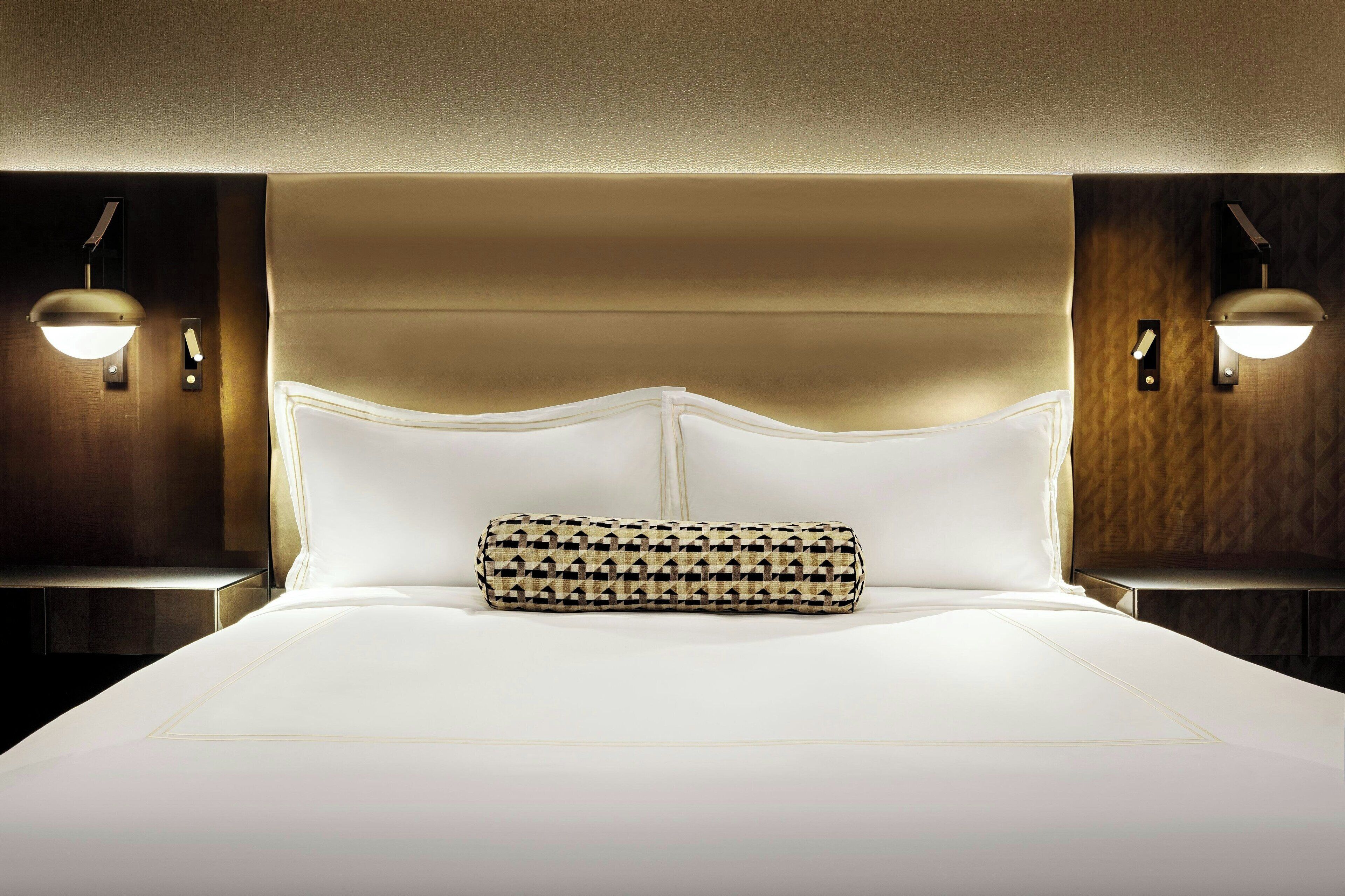 A hotel room with a large bed, a headboard, two bedside lamps, and a patterned throw pillow.
