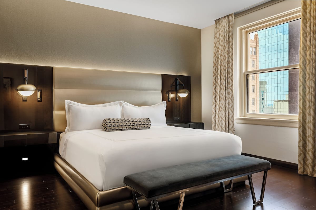 A modern hotel room with a large bed, bedside lamps, a bench, and a window with curtains.