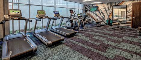 Un gimnasio con cintas de correr, elípticas y bicicletas estáticas, ventanas amplias y una alfombra estampada.