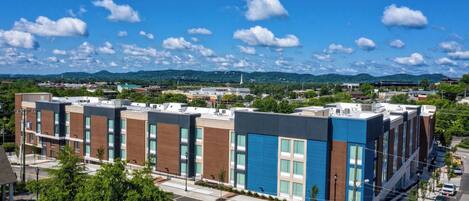 A modern multi-story building with a mix of brick and glass facades, located in a suburban area with a clear sky and distant hills.