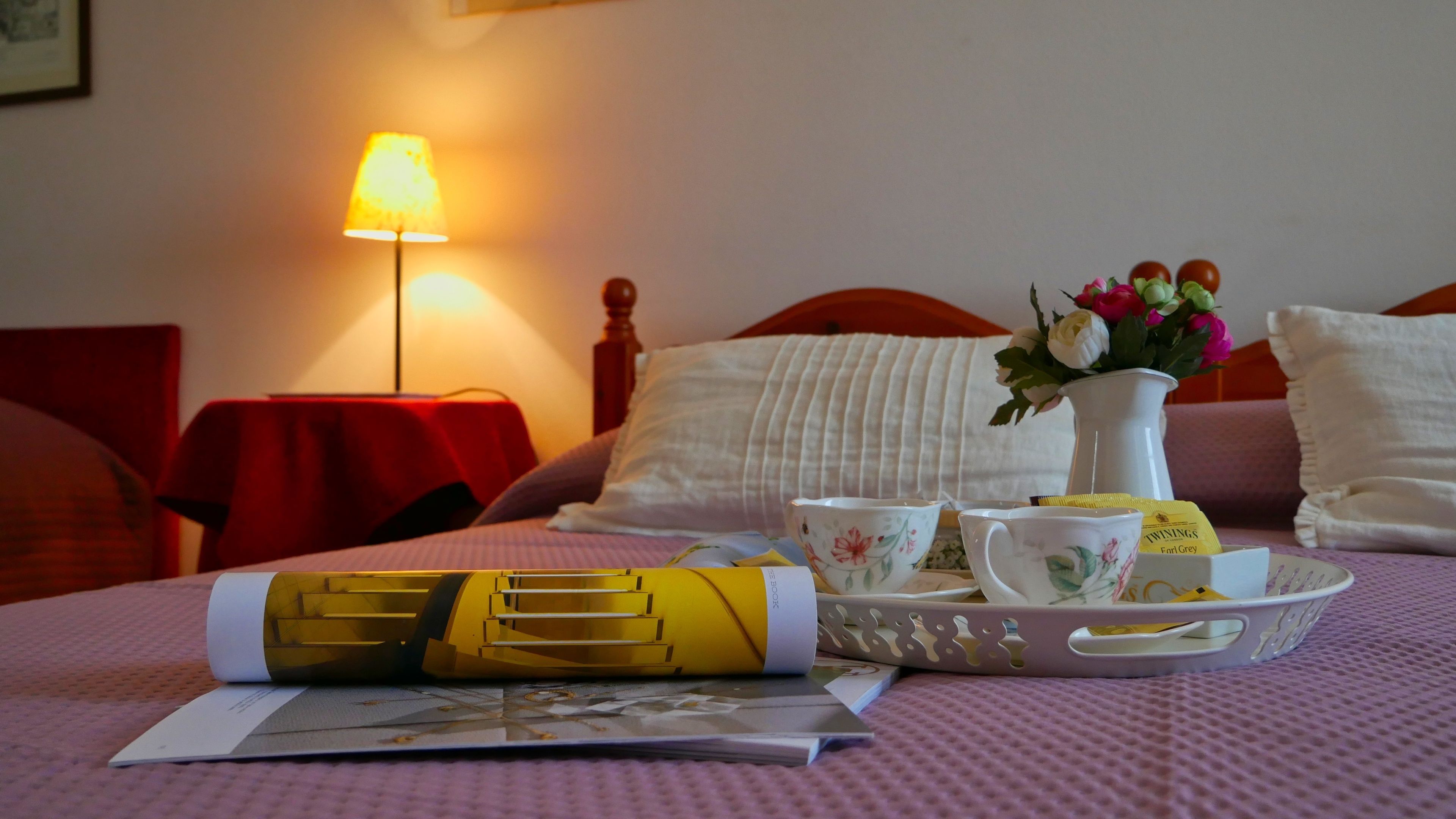 A neatly made bed with a floral-patterned pillow, a bedside table with a lamp, and a tray with teacups and a vase of flowers.