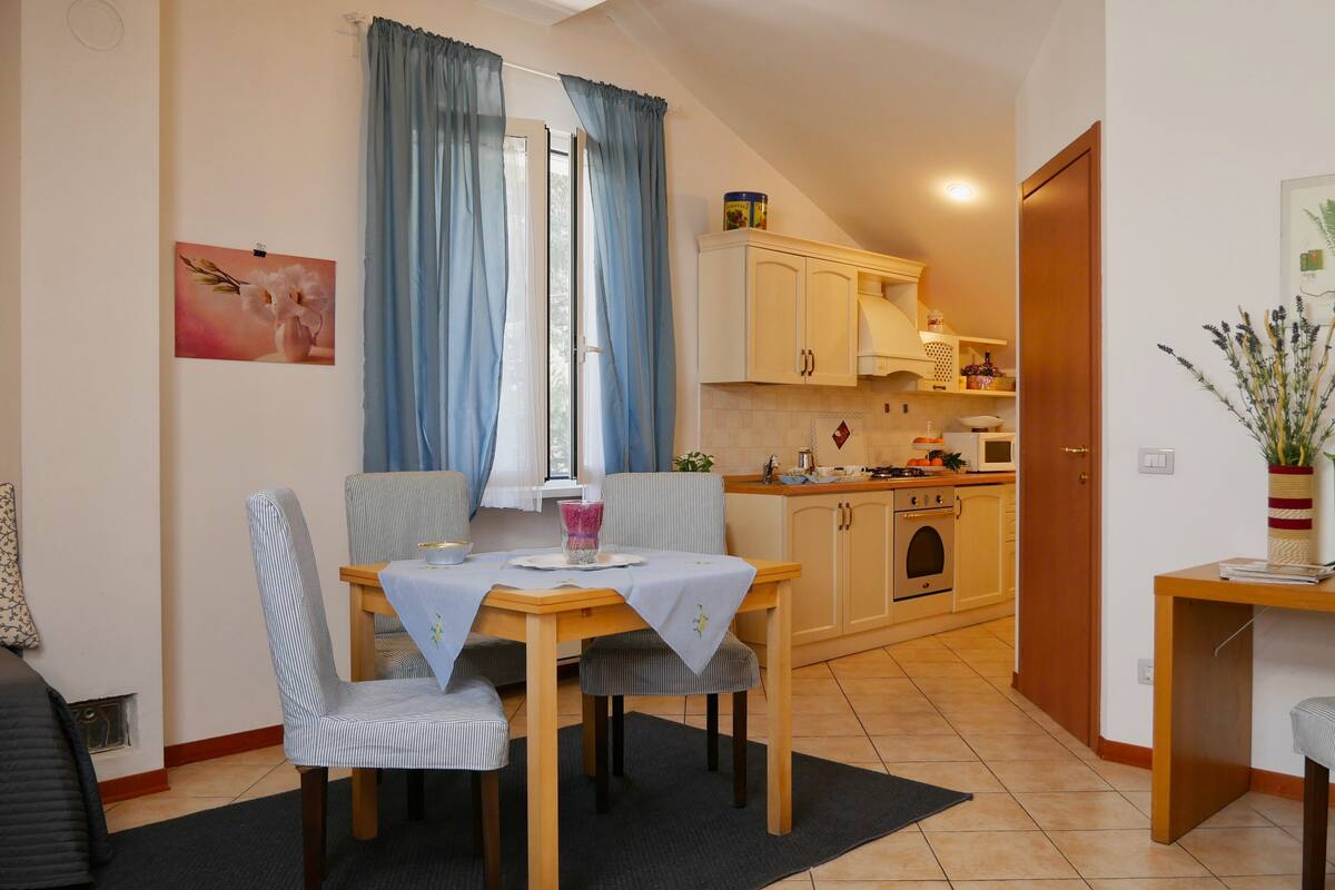 A dining area with a table and chairs, a kitchen in the background, and a door leading to another room.