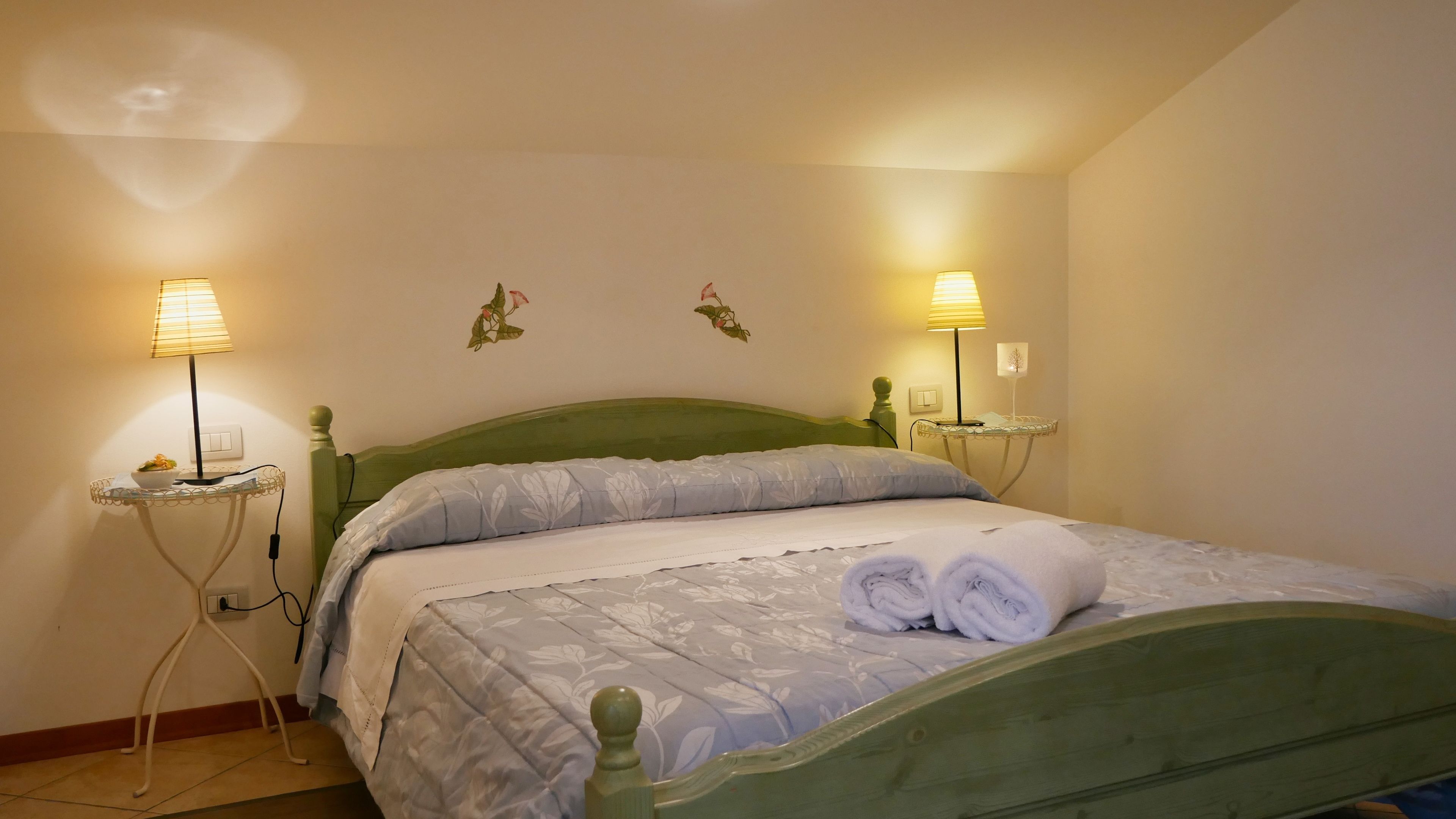 A neatly made bed with a floral bedspread, two bedside lamps, and a small table with a bowl of fruit.