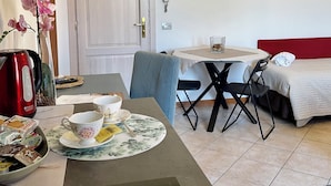 A compact hotel room with a dining area, a bed with a red headboard, and a small table with a kettle and breakfast items.