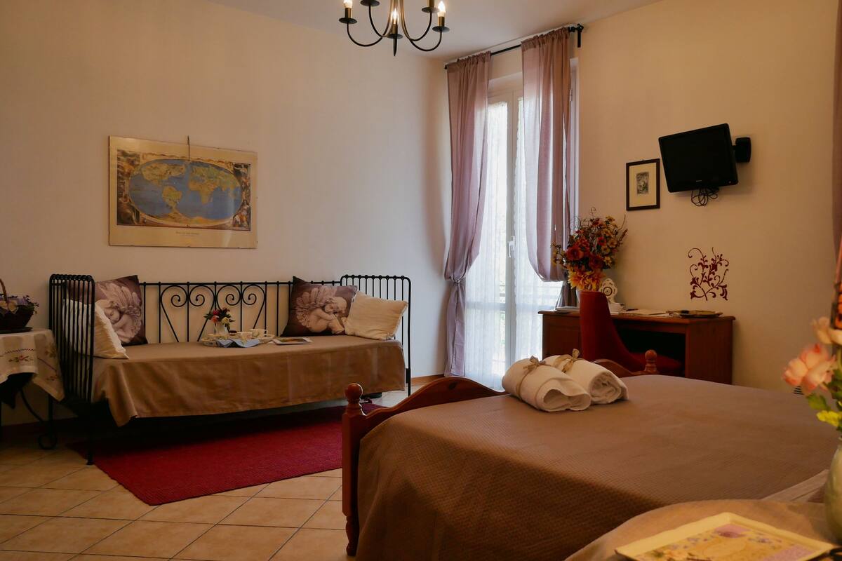 A hotel room with a bed, a smaller bed, a desk with a computer, a TV, a chandelier, and a window with curtains.
