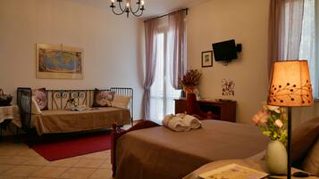 A hotel room with a bed, a smaller bed, a desk with a computer, a TV, a chandelier, and a window with curtains.