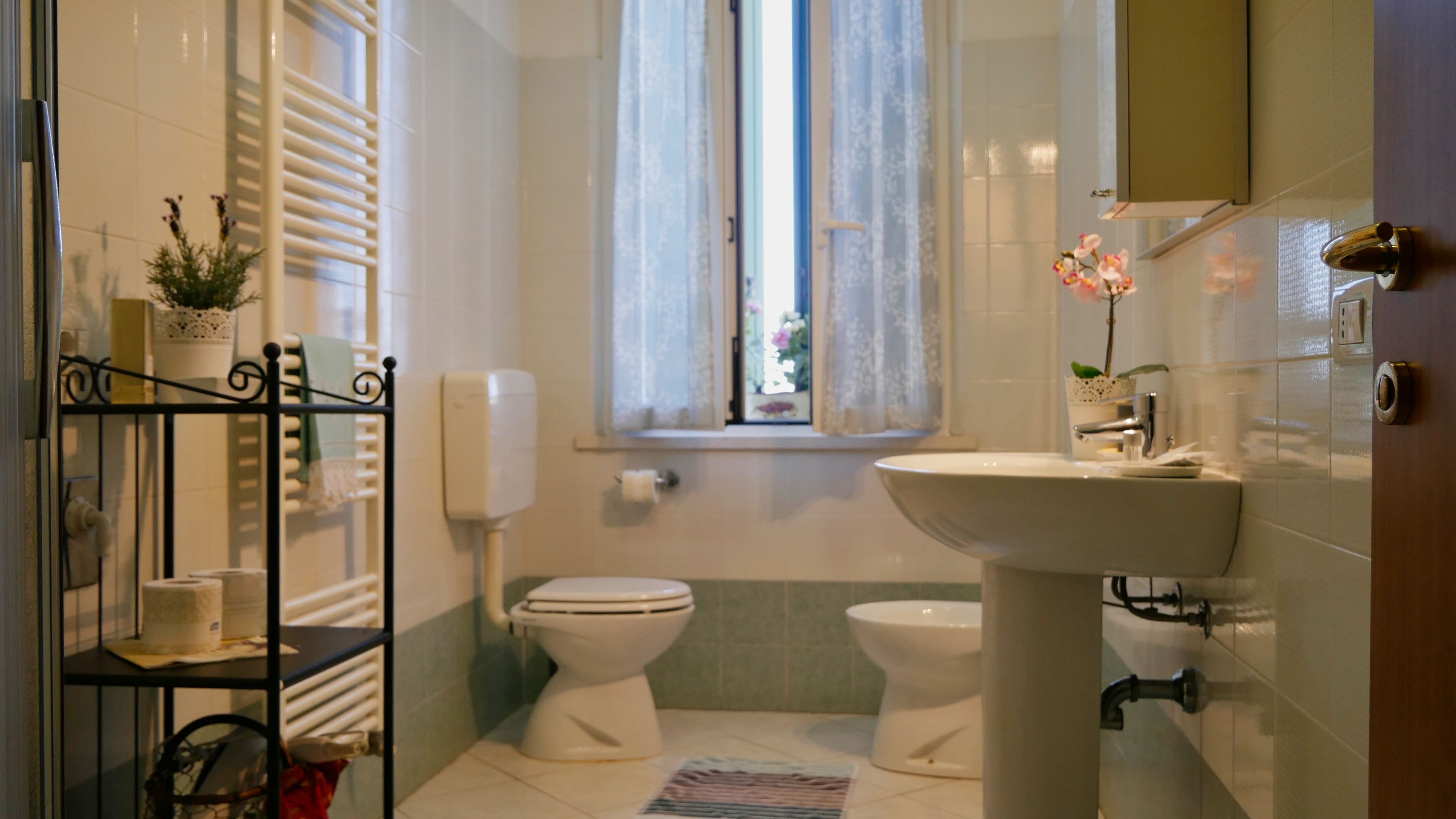 A bathroom with two toilets, a sink, a window with lace curtains, and a shelf with toiletries.