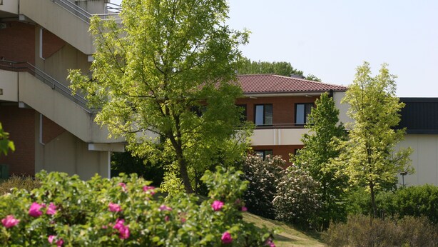 A multi-story building with a red-tiled roof, surrounded by lush greenery and flowering bushes. - Sportpark Hotel (Bitburg)
