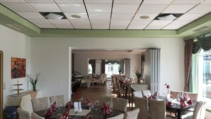 A dining area with multiple tables and chairs, a buffet setup, and a view into another room with a dining table and chairs. - Sportpark Hotel (Bitburg)