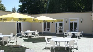 An outdoor patio area with tables and chairs under yellow umbrellas, a brick floor, and a building with windows and doors. - Sportpark Hotel (Bitburg)