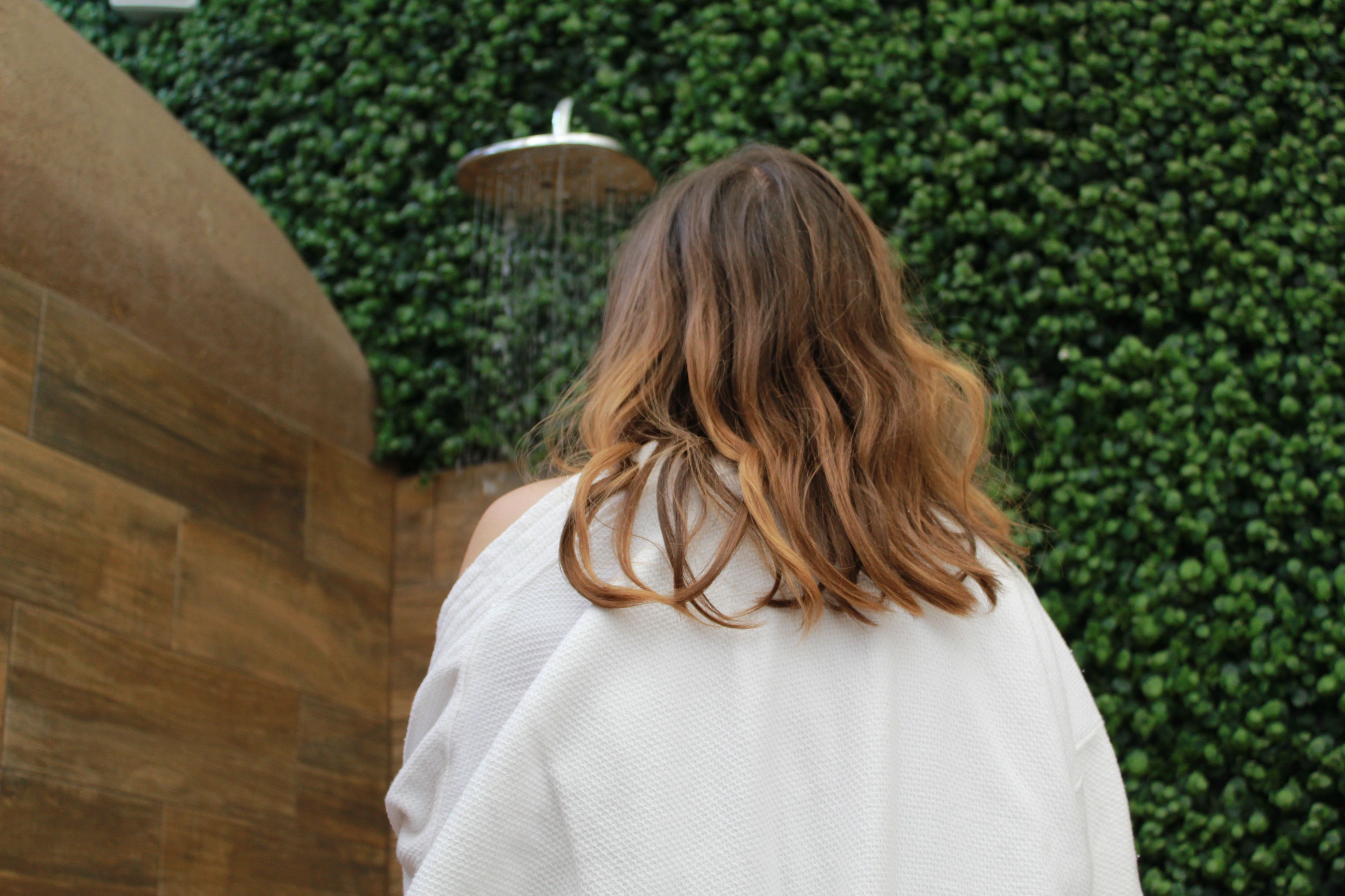 A person with long, wavy hair standing in front of a shower with a green wall and a wooden structure to the left.