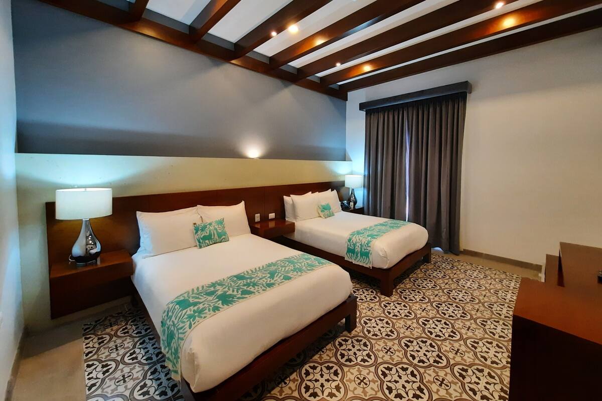 Une chambre d’hôtel avec deux lits, un sol à motifs, des poutres en bois au plafond et une grande fenêtre avec des rideaux.