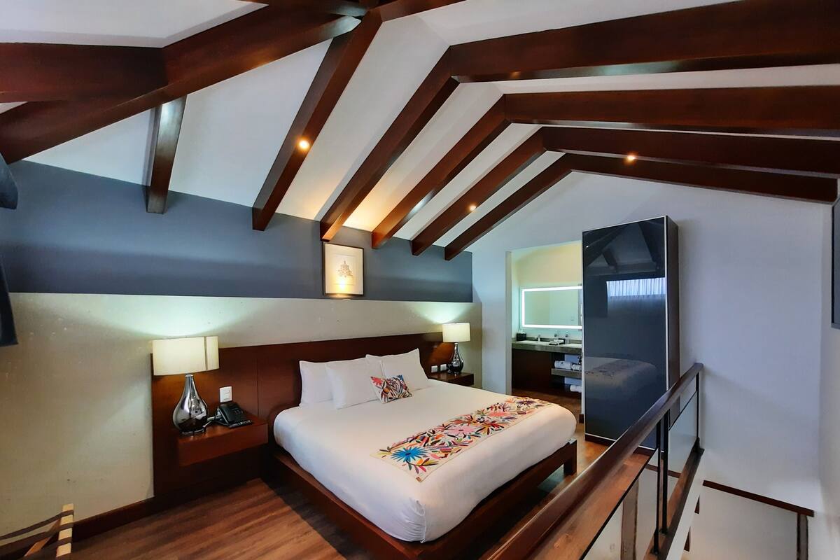 Une chambre d’hôtel avec un plafond en bois, un lit recouvert d’une couverture colorée, une table de chevet avec une lampe, et une salle de bain visible à travers une porte ouverte.