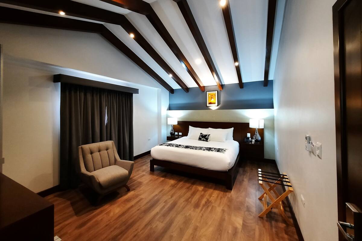 Une chambre d’hôtel moderne dotée d’un grand lit, d’une chaise, d’un bureau et d’un plafond avec des poutres apparentes et un éclairage encastré.