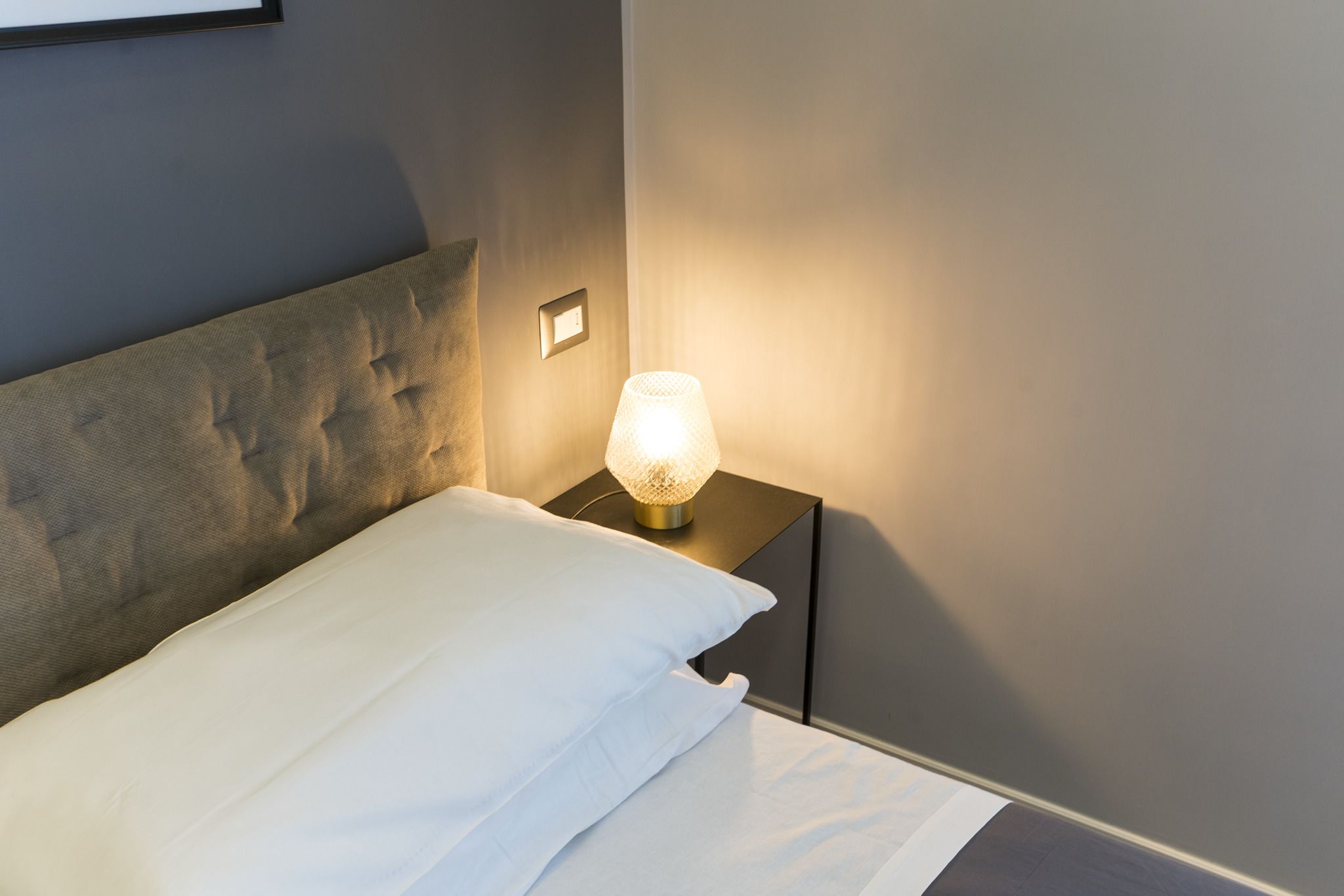 A neatly made bed with a headboard, a bedside lamp, and a power outlet on the nightstand.