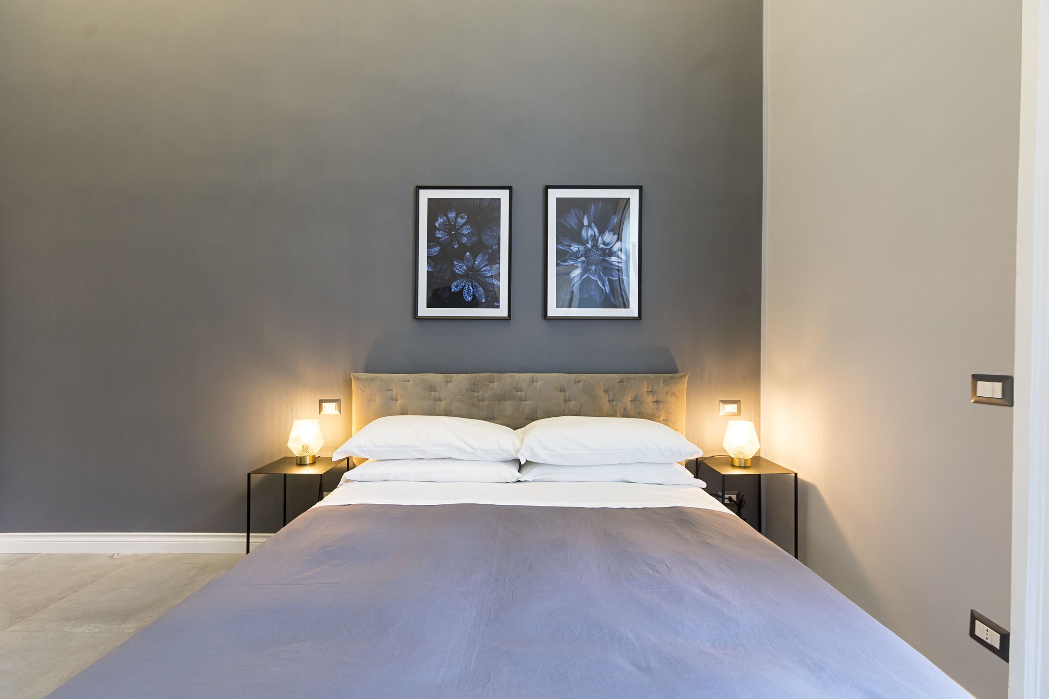 A bedroom with a large bed, two bedside lamps, and two framed floral artworks on the wall.