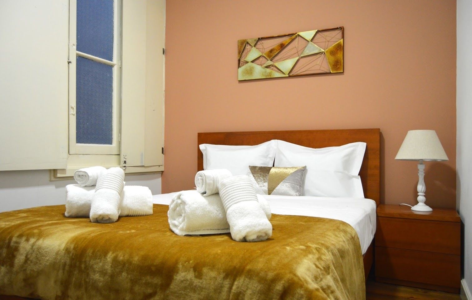 A hotel room with a bed, pillows, a bedside table with a lamp, and a window with curtains.