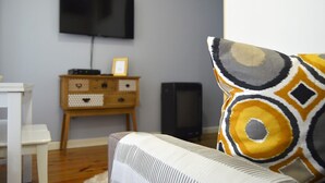 En stue med et fladskærms-tv monteret på væggen, et træskab med skuffer og en mønstret pude på en hvid sofa.