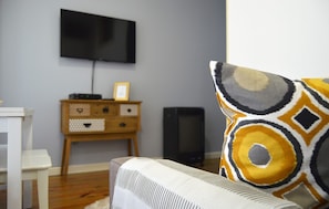 Un salon avec un téléviseur à écran plat fixé au mur, un meuble en bois avec des tiroirs et un coussin décoratif à motifs sur un canapé blanc.