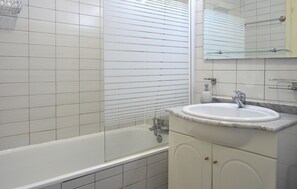Une salle de bain avec des murs recouverts de carreaux blancs, une baignoire blanche, un lavabo blanc avec un plan de toilette en marbre et une cabine de douche en verre.