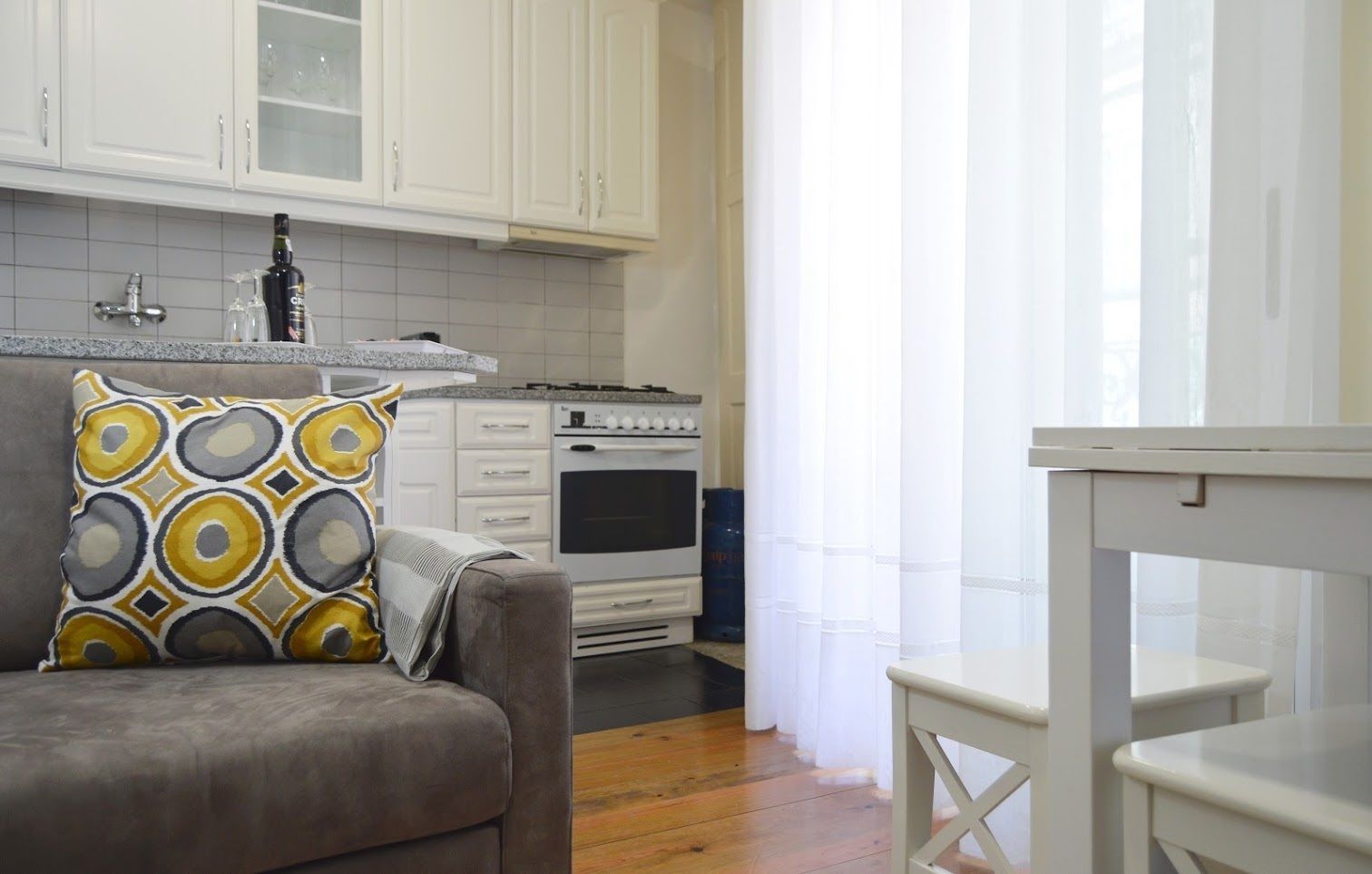 A kitchen with white cabinets, a stove, and a sink. There is a gray sofa with a patterned pillow and a white table.