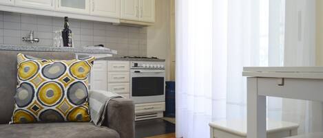 A kitchen with white cabinets, a stove, and a sink. There is a gray sofa with a patterned pillow and a white table.
