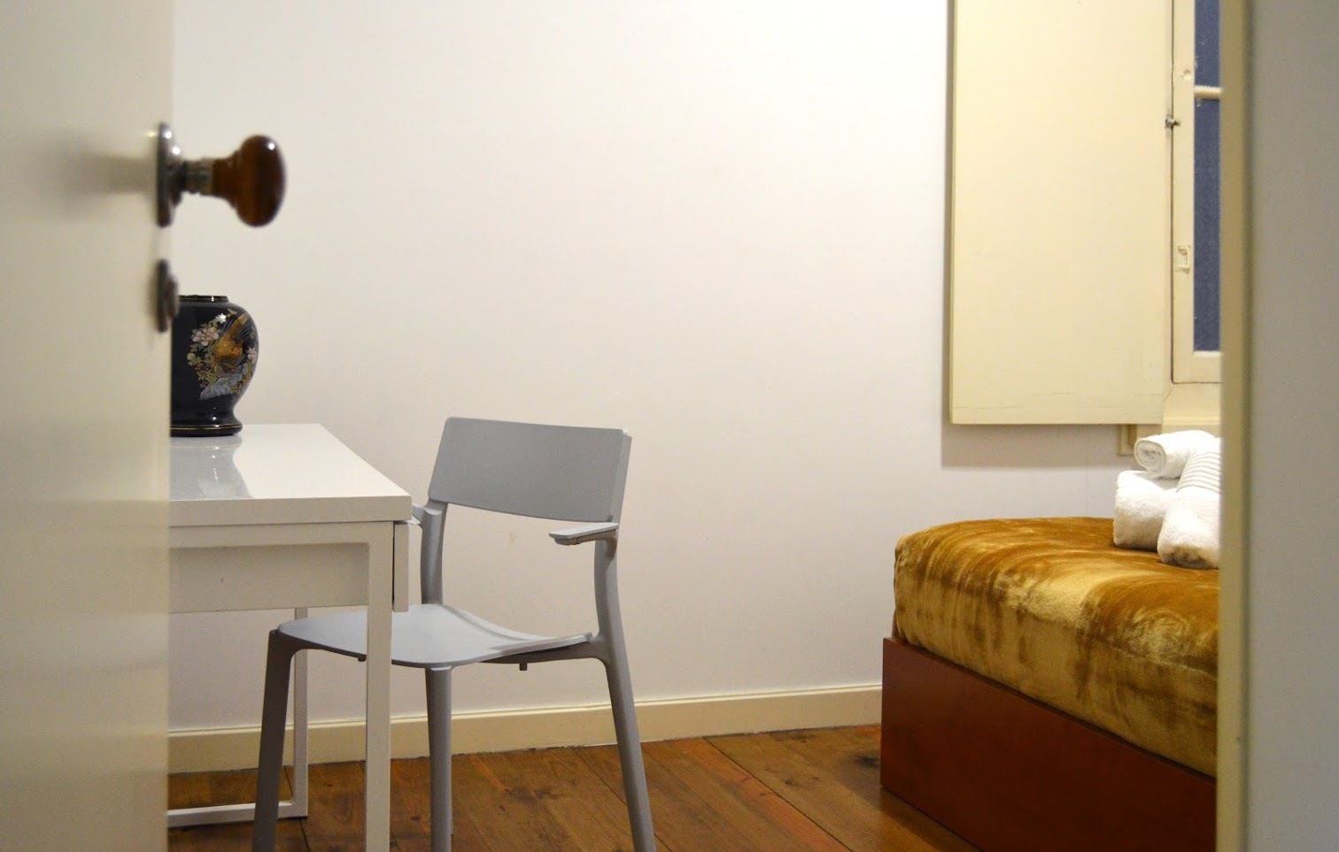 Ein Zimmer mit einem weißen Schreibtisch, einem grauen Stuhl, einem Bett mit brauner Kopfteil und einer weißen Wand.