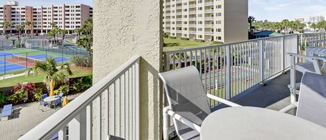 A balcony with outdoor seating, a tennis court, and a view of a multi-story building.